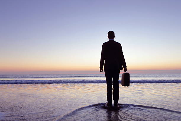 Businessman stands in sea shallows, sunset, Cornwall Businessman stands in sea shallows, sunset, Cornwall